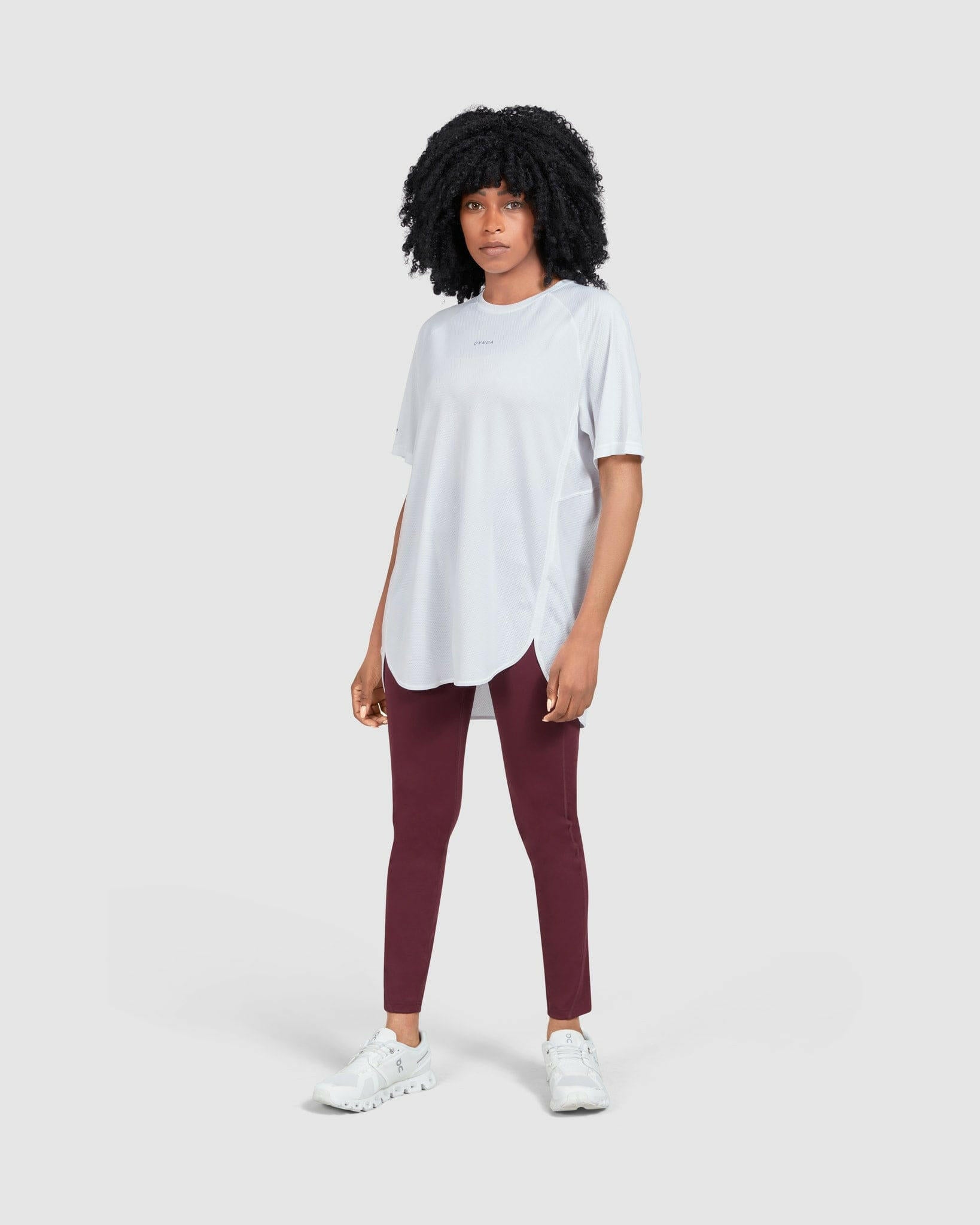 A young woman in a casual short-sleeved white BREATHE T-SHIRT with qynda logo and maroon leggings standing against a white background, wearing white sneakers, embodying a simple, sporty, and comfortable style.