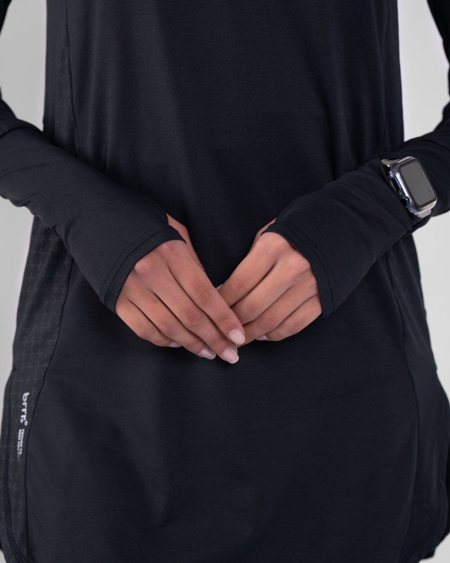 A close-up photo of a model in a black high neck long sleeve shirt ARMA by Qynda with cooling fabric technology, holding their hands together at the lower, showing a smartwatch on their left wrist.