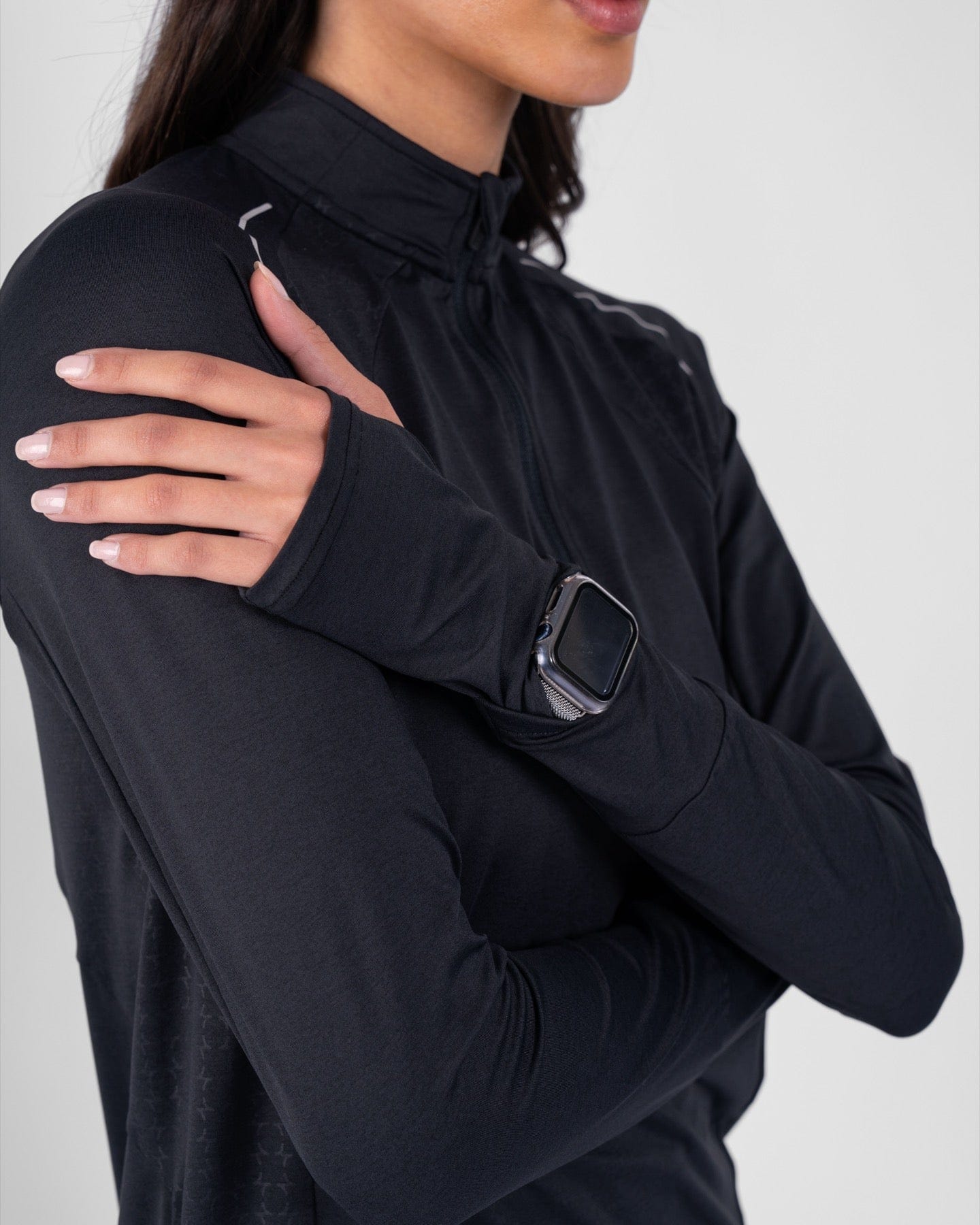 Close-up of a model wearing black high neck long sleeve shirt ARMA by Qynda with moisture-resistant fabric technology, sporting a smartwatch on her wrist, hand resting on her shoulder, against a grey background.