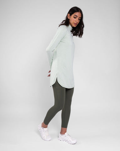 A model posing wearing a high neck long sleeve shirt ARMA by Qynda and olive green leggings paired with white sneakers, against a light gray background.