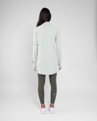 A model seen from behind wearing a high neck long sleeve shirt ARMA by Qynda and olive green leggings paired with white sneakers, against a light gray background.
