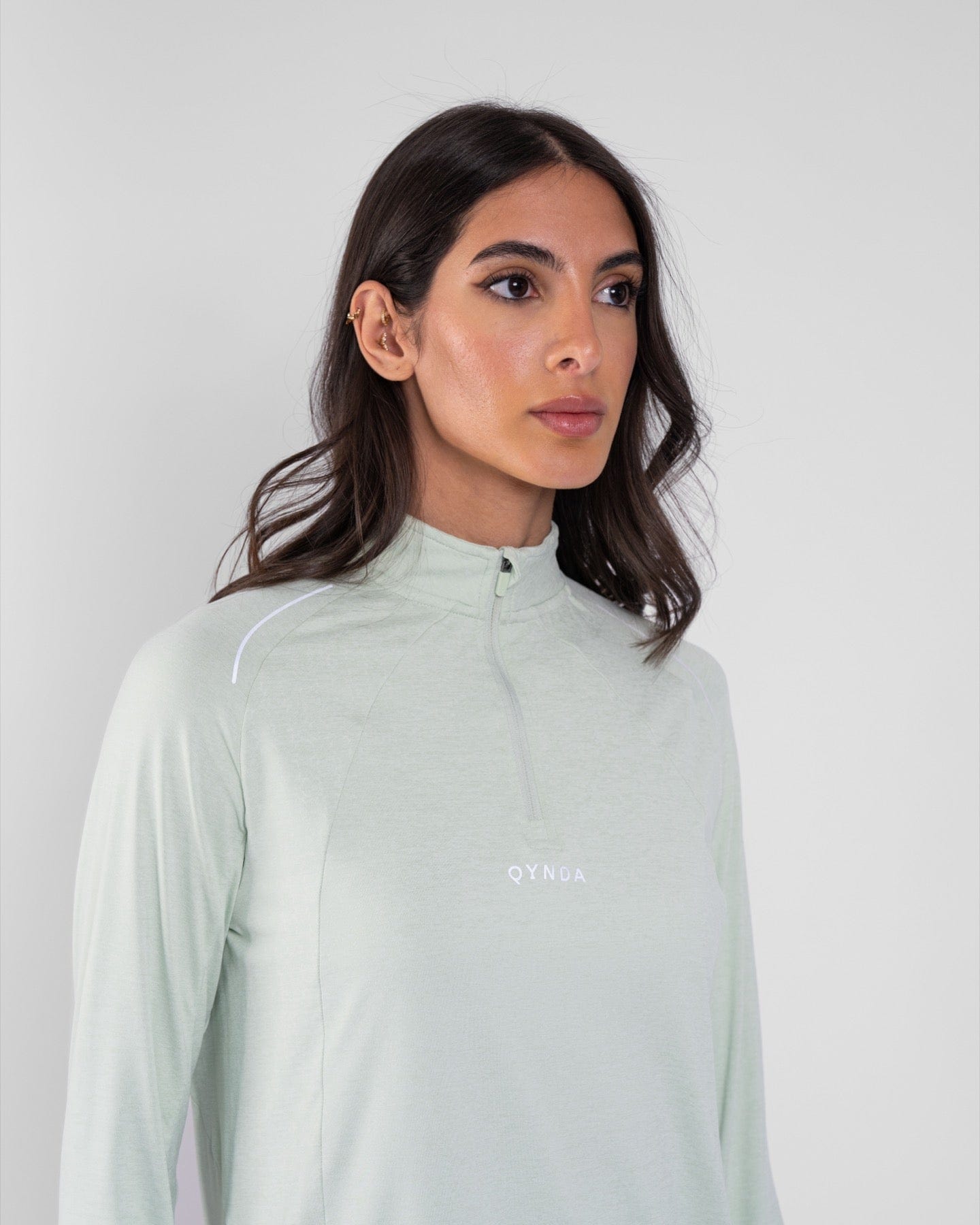 A model with medium-length brown hair wearing a high neck long sleeve shirt ARMA by Qynda featuring moisture-resistant technology, slightly turned to her left, against a light gray background.