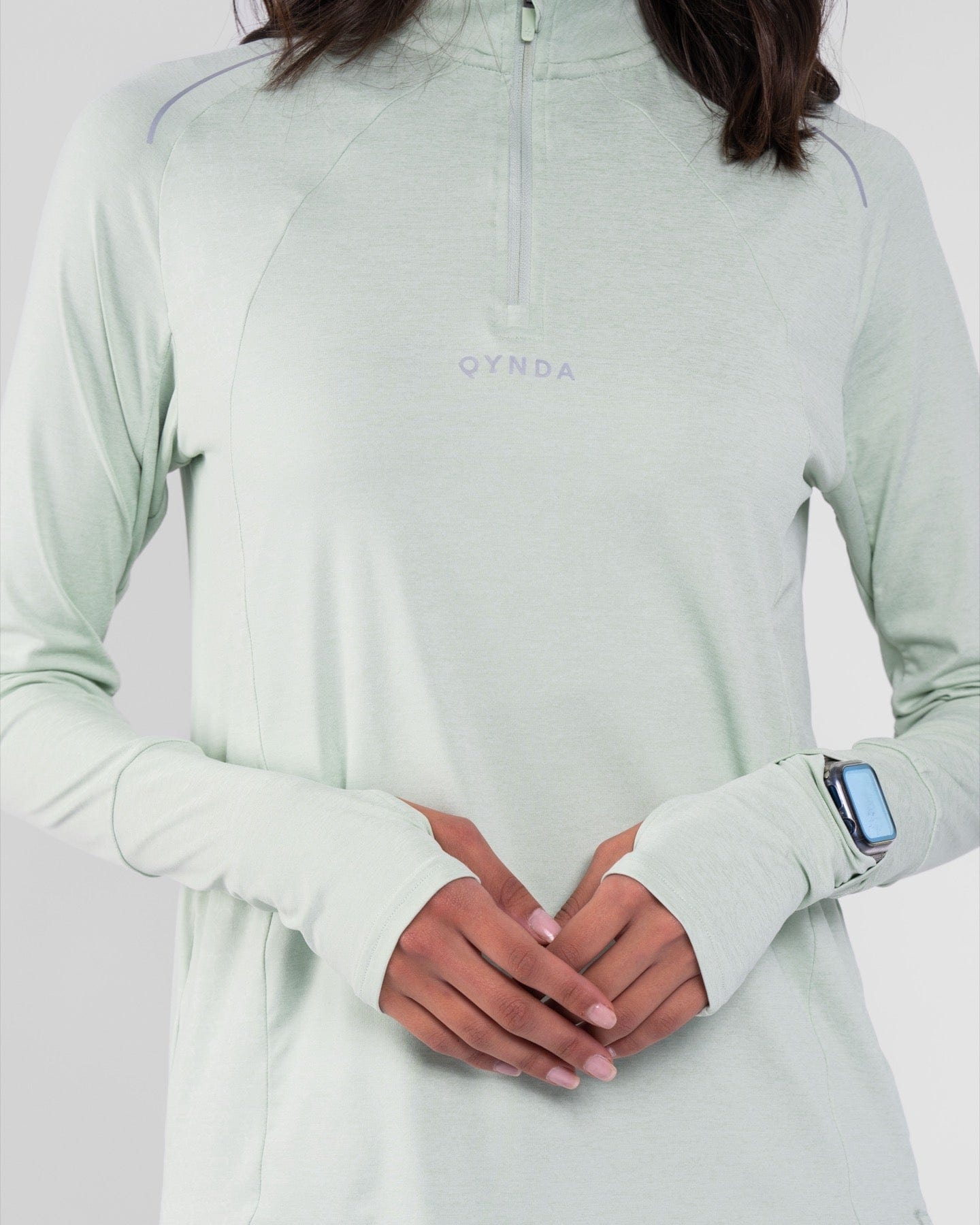 A close-up photo of a model in a Sage high neck long sleeve shirt ARMA by Qynda with cooling fabric technology, holding their hands together at the lower, showing a smartwatch on their left wrist.