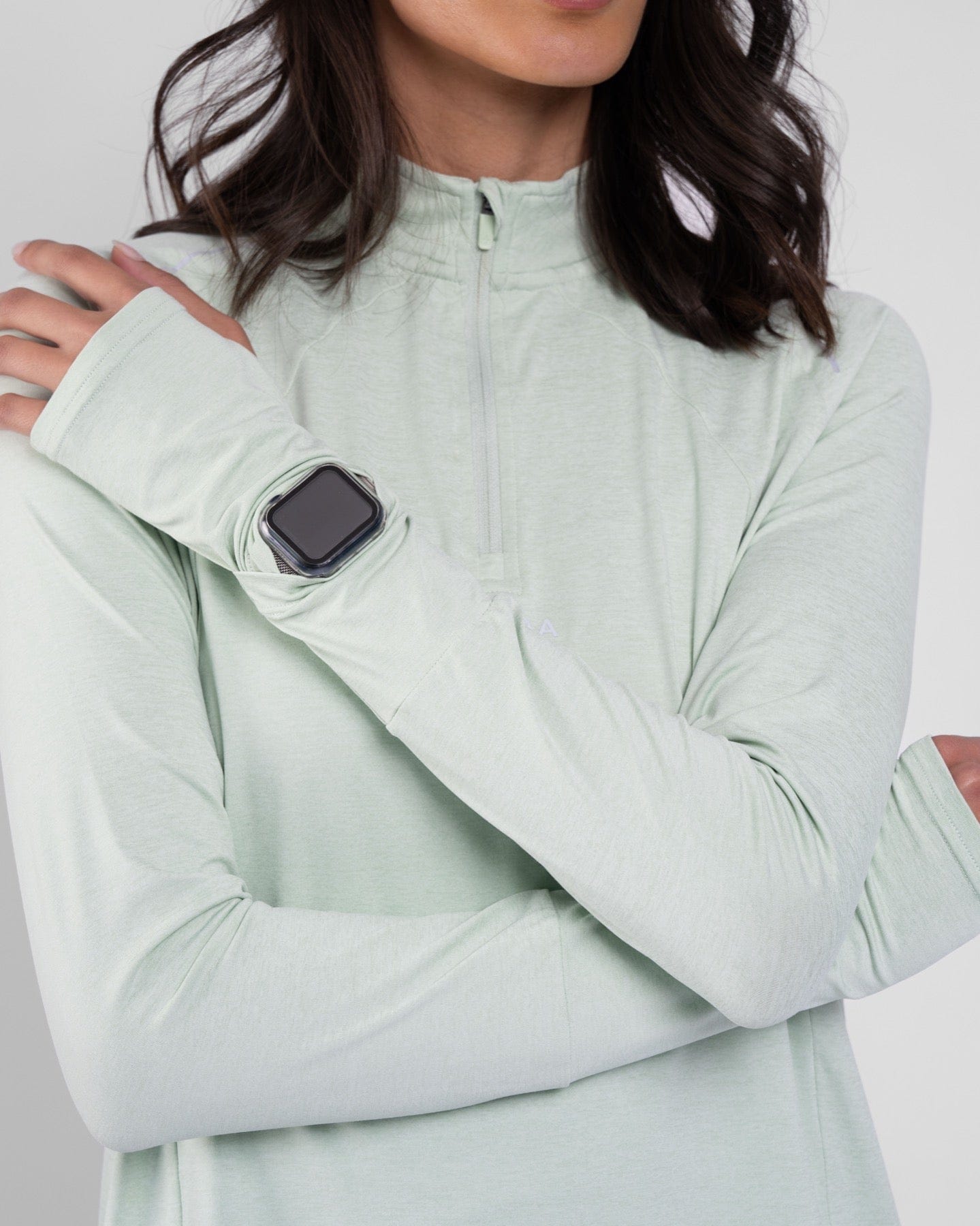 Close-up of a model wearing Sage high neck long sleeve shirt ARMA by Qynda with moisture-resistant fabric technology, sporting a smartwatch on her wrist, hand resting on her shoulder, against a grey background.