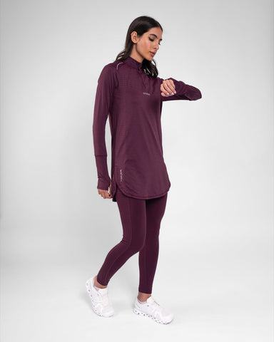 A woman in a maroon high neck long sleeve shirt ARMA by Qynda, featuring quick-drying fabric, stands against a grey background, looking to her right.