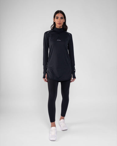 A model wearing a modest sleek black long-sleeve ARMA top by Qynda with cooling fabric technology and leggings, standing against a light gray background, facing the camera with a relaxed pose.
