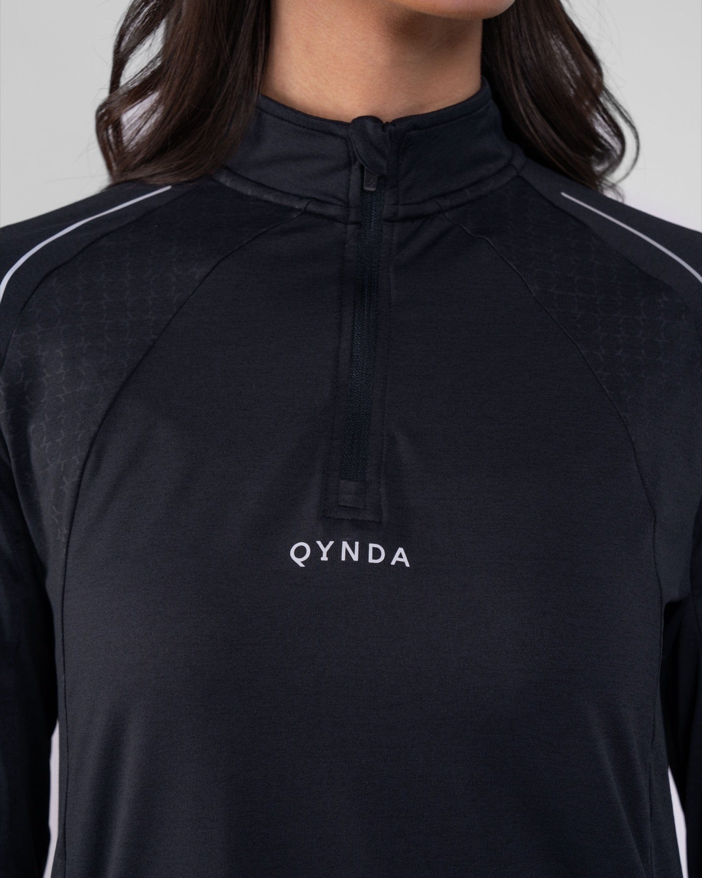 Close-up of a woman wearing a black, high neck long sleeve shirt ARMA  by Qynda with a subtle hexagonal pattern, featuring a half-zip.